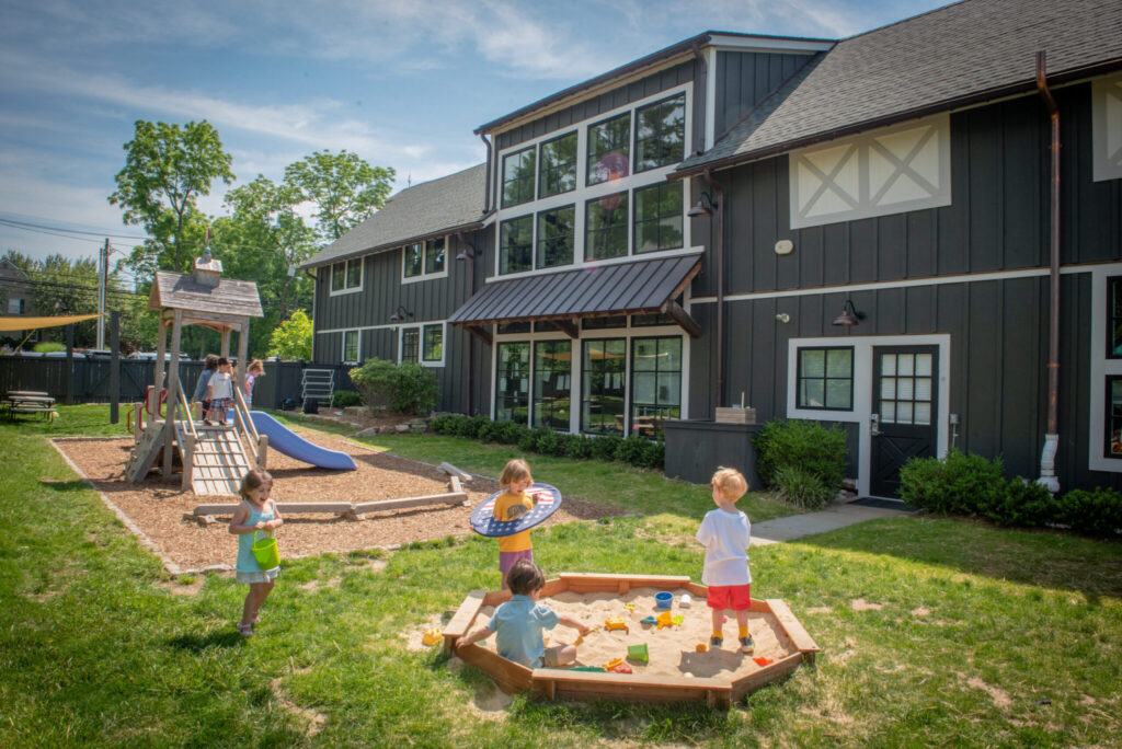 Children playing outside at Haven Gladstone Playground in New Jersey, featuring a wooden play structure, sandbox, and modern preschool building under a sunny blue sky.