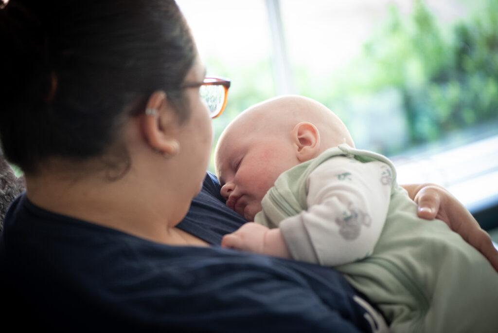 A baby in a green onesie is asleep in the arms of a woman.